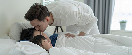 Un homme debout embrasse son mari sur le front avant d’aller travailler, tandis que leur enfant dort.