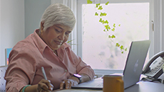 Une femme d’âge moyen est assise à un bureau, devant un ordinateur portable ouvert, et elle écrit dans un bloc-notes.