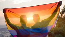 Two people holding a Pride flag are silhouetted by a glowing sunset.