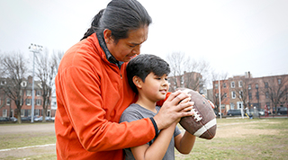 An Indigenous father teaches his son how to throw a football outdoors in a park. 
