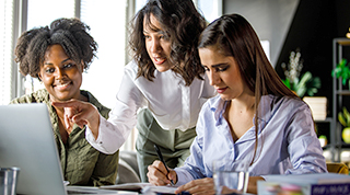 Three racially diverse female TD colleagues have a discussion while seated around a laptop in an office. 