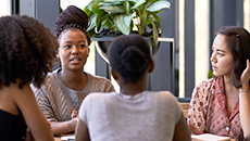 A group of racially diverse women seated around a table hold a discussion.  