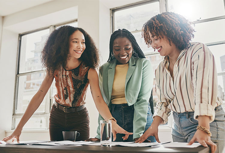 Three Black women entrepreneurs look at financial documents spread out over a table in a sunny office. 