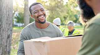 Two Black volunteers carry a box of supplies at a volunteer event in a park. 