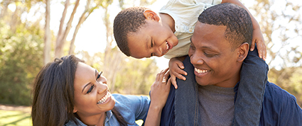 A Black family smiles together in a park as a child sits on their father’s shoulders. 