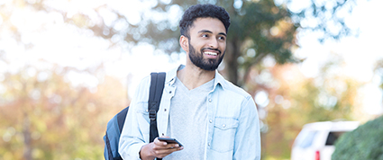 A South Asian student carrying a backpack checks his phone while walking to a university class.