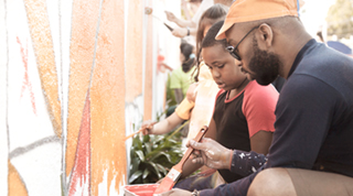 A Black father and son participate in painting a colourful mural on the side of a building.