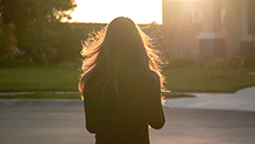 A woman stands silhouetted from behind as the sun sets over a house on a suburban street.