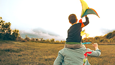 A child holds a kite while seated on their parent’s shoulders in a field.