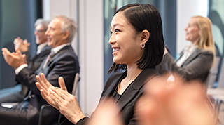 An Asian female executive applauds while seated in a room of her colleagues at a conference.