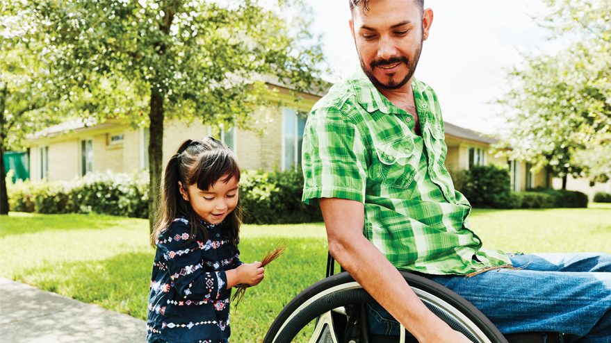 Father in wheelchair on sidewalk with daughter walking behind