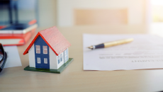 A wooden toy house on a table beside a piece of paper and pen
