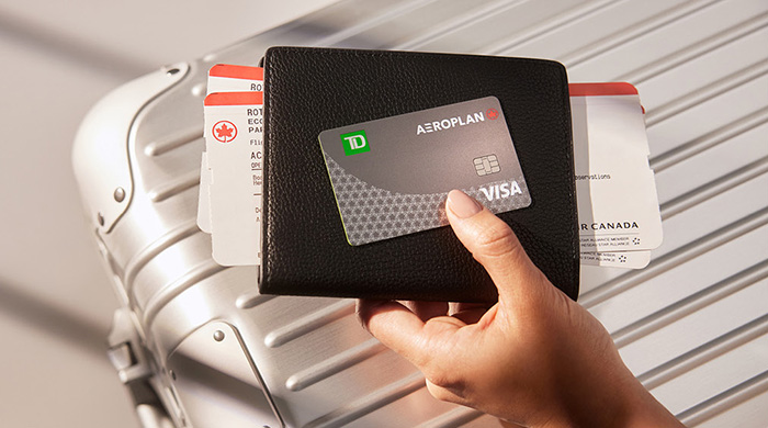 Une main tenant un étui à passeport, une carte d’embarquement d’avion et une carte Visa TD Aéroplan, avec une valise en arrière-plan