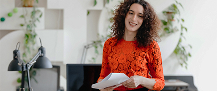Une propriétaire de PME aux cheveux longs et portant un chandail orange vif sourit tout en examinant des documents à son bureau.