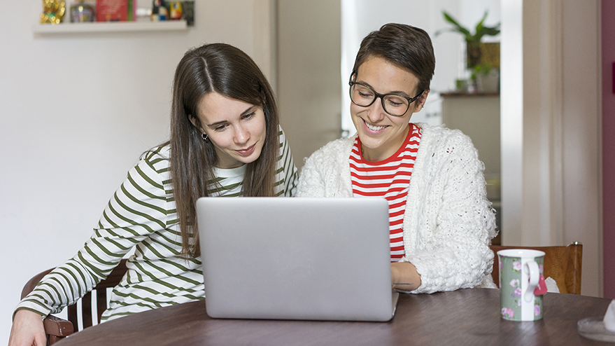 Two girlfriends at home sitting at table, using laptop