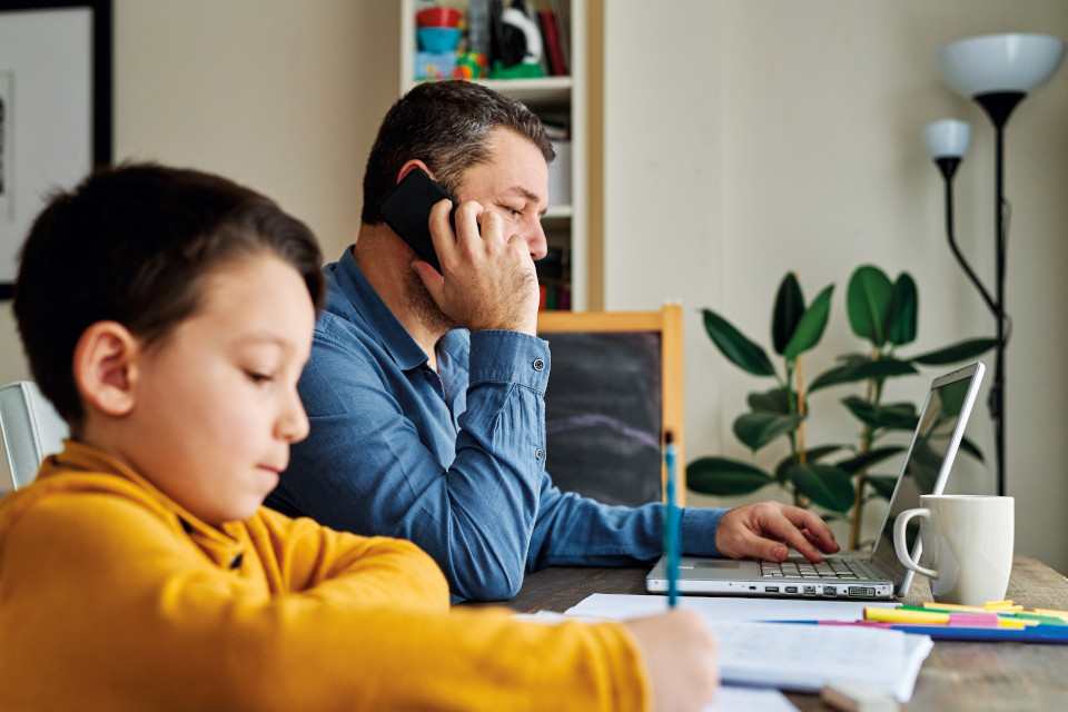 Un homme travaille à la maison sur son ordinateur portable et son enfant fait ses devoirs assis à ses côtés