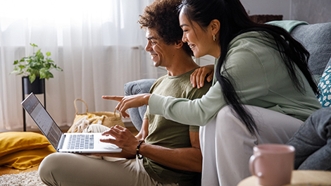 Copy space shot of young man sitting on the floor and using laptop, organizing finances and online banking. His girlfriend is sitting on the sofa, behind him, and pointing at something on the screen.