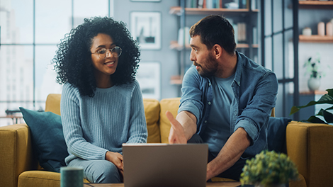 Diverse Multiethnic Couple are Sitting on a Couch Sofa in Stylish Living Room and Choosing Items to Buy Online with Laptop Computer. Friends or Colleagues are Discuss Work Projects.